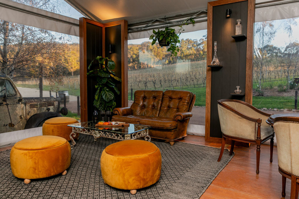 A rustic indoor lounge area with a brown leather sofa, two mustard yellow velvet ottomans, a glass-top coffee table, and cream upholstered chairs, set against large windows overlooking a vineyard.
