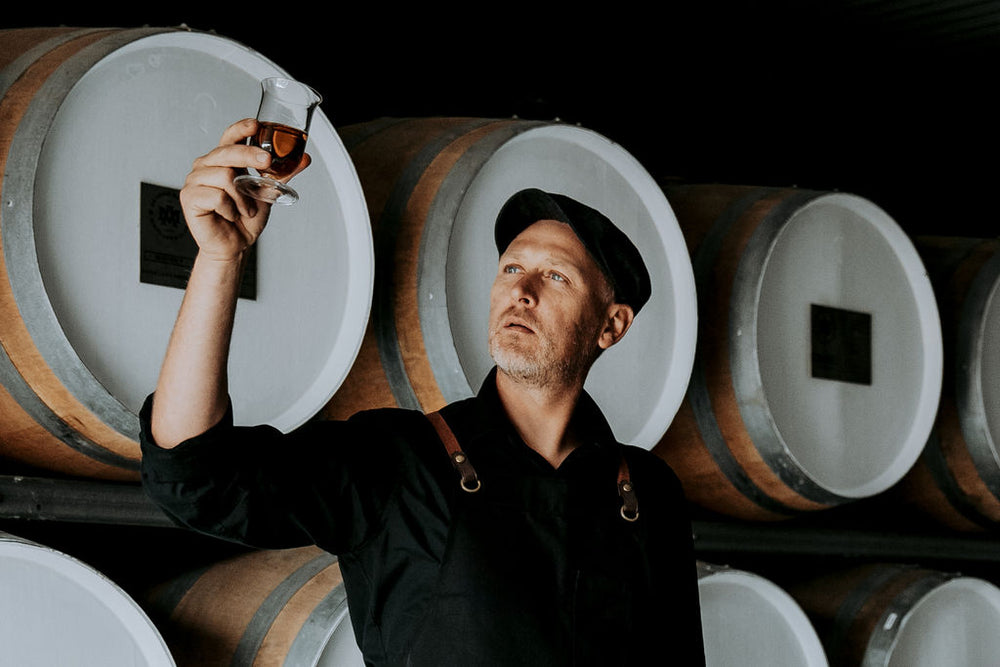 A man in a black shirt and cap holds up a small glass of amber liquid in a wine or whiskey cellar, with large wooden barrels visible in the background.