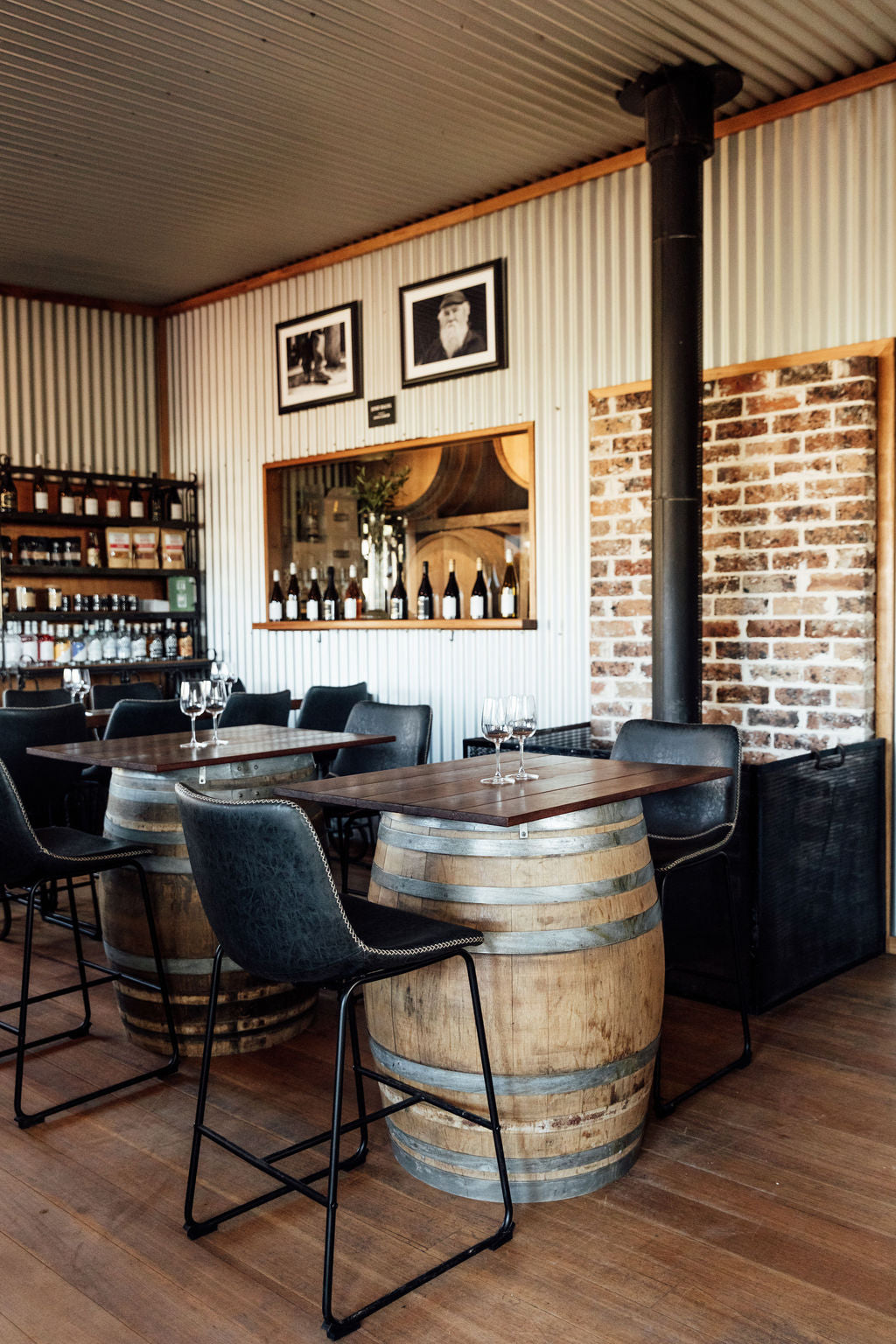 A rustic indoor bar or restaurant setting with wooden barrel-height tables, black bar stools, a corrugated metal wall, brick wall sections, and a wine display in the background.